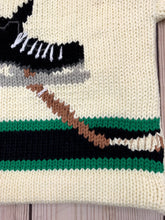 ivory long sleeve sweater with black band on waist and arms with green trim and hockey skate/stick in center